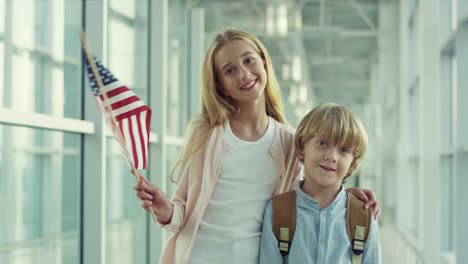 Portrait-Shot-Of-The-Happy-Kids,-Girl-And-Boy,-Standing-In-The-Airport-And-Smiling-To-The-Camera-With-American-Flag