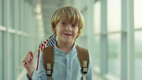 Close-Up-Of-The-Cute-Small-Boy-Standing-In-The-Airport-Passage,-Looking-To-The-Camera-And-Waving-With-American-Flag