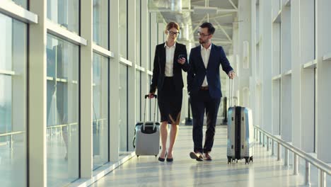 Attractive-Man-And-Woman-In-Business-Style-Walking-The-Corridor-Of-The-Airport-With-Suitcases,-Speaking-And-Woman-Tapping-On-The-Smartphone