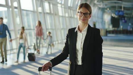 Portrait-Of-The-Beautiful-Businesswoman-In-Glasses-Standing-In-The-Airport-With-Suitcase-And-Smiling-To-The-Camera-In-The-Airport