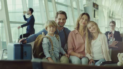 Portrait-Shot-Of-The-Cute-Happy-Family-With-Son-And-Daughter-Sitting-In-The-Airport-With-Suitcases-And-Smiling-To-The-Camera-While-Waiting-For-Their-Departure