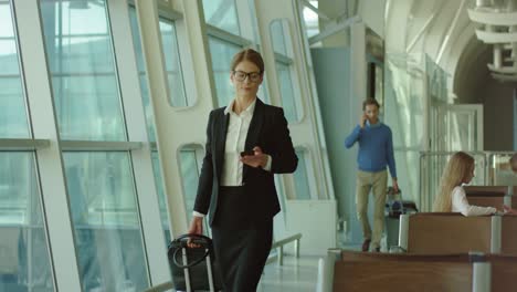Pretty-Businesswoman-In-Glasses-Walking-The-Airport-Hall-With-A-Smartphone-In-Hands-And-Then-Sitting-On-The-Bench