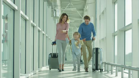 Nice-Cheerful-Mother,-Father-And-Son-Going-Through-The-Passage-In-The-Airport-With-Suitcases-On-The-Wheels-To-The-Plane-As-Having-Vacations-Tour