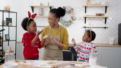 Cheerful-Woman-In-Apron-With-Little-Kids-Standing-At-Table-In-Home-Christmassy-Decorated-Kitchen-And-Having-Fun-While-Making-Xmas-Cookies