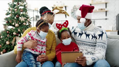 Family-In-Masks-With-Kids-Gathered-At-Home-Near-Christmas-Tree-On-Holidays-Using-Electronic-Device