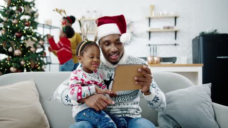 Close-Up-Of-Happy-Little-Kid-Sitting-With-Dad-In-Santa-Hat-At-Decorated-Room-And-Watching-Cartoons-On-Tablet