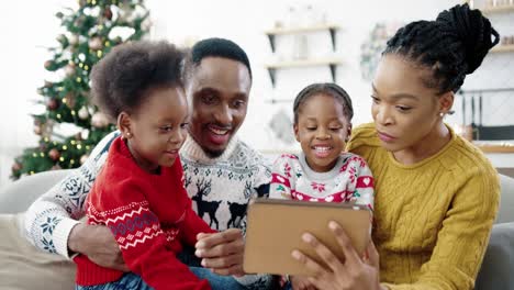 Close-Up-Of-Happy-Family-At-Home-Sitting-In-Decorated-Room-Near-Christmas-Tree-And-Using-Tablet-Small-Cute-Kids-Sitting-With-Mom-And-Dad-And-Tapping-On-Tablet-Choosing-Presents-Online