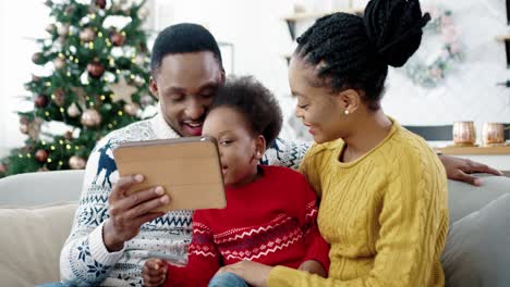 Portrait-Of-Happy-Little-Kid-Sitting-On-Sofa-In-Decorated-Room-Near-New-Year-Tree-With-Mom-And-Dad-And-Tapping-On-Tablet-While-Playing