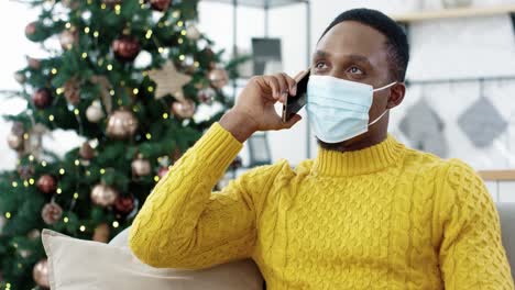 Close-Up-Portrait-Of-Cheerful-Guy-In-Medical-Mask-Sitting-On-Sofa-In-Decorated-Room-At-Home-Near-Christmas-Tree-And-Chatting-On-Smartphone