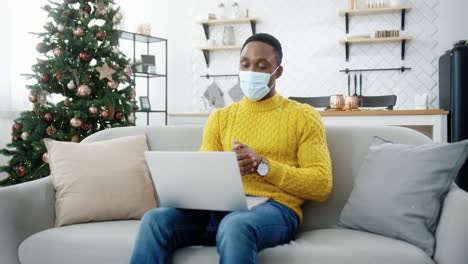 Handsome-Guy-In-Medical-Mask-Sitting-On-Couch-In-Decorated-Room-At-Home-Near-Christmas-Tree-With-Twinkle-Lights-And-Video-Chatting-On-Laptop