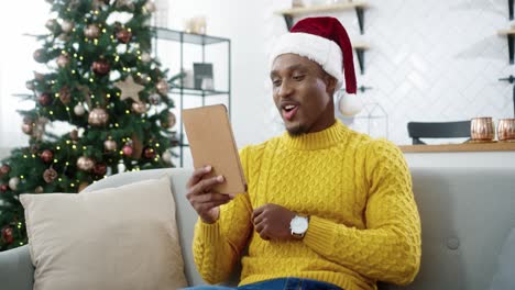 Portrait-Of-Cheerful-Young-Male-Wearing-Santa-Hat-Watching-Video-Online-On-Tablet-And-Laughing-Indoors-In-Room-With-Pretty-Christmas-Decorated-Tree