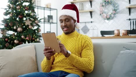 Primer-Plano-De-Un-Comprador-Masculino-Feliz-Con-Sombrero-De-Santa-Sentado-En-Una-Casa-Decorada-Cerca-De-Un-árbol-Brillante-De-Navidad-Y-Comprando-En-Línea-En-Una-Tableta-Pagando-Con-Tarjeta-De-Crédito-En-Las-Ventas-De-Navidad