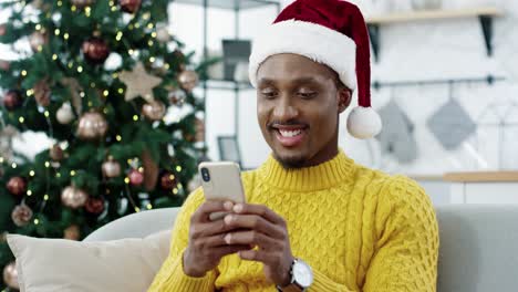 Nahaufnahme-Eines-Gutaussehenden-Mannes,-Der-Auf-Einem-Sofa-In-Einem-Modernen-Zuhause-In-Der-Nähe-Eines-Glühenden-Weihnachtsbaums-Sitzt-Und-Auf-Dem-Smartphone-SMS-Schreibt-Und-Urlaubsgrüße-An-Freunde-Sendet