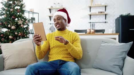 Glücklicher-Gutaussehender-Mann-Mit-Weihnachtsmütze-Videochattet-Auf-Tablette,-Während-Er-Zu-Hause-In-Der-Nähe-Des-Geschmückten-Neujahrsbaums-Sitzt
