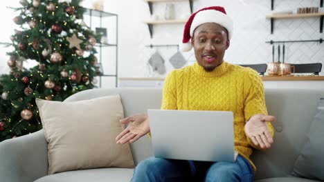 Cheerful-Handsome-Guy-In-Xmas-Hat-Videochatting-On-Computer-While-Sit-Near-Decorated-Glowing-New-Year-Tree-At-Home