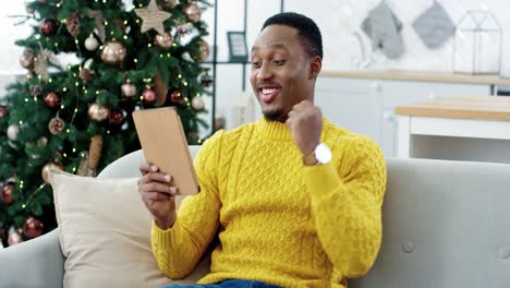 Portrait-Of-Joyful-Man-Buyer-Sitting-In-Decorated-Home-Near-Christmas-Tree-With-Surprised-Face-And-Buying-Online-On-Tablet-Payment-With-Credit-Card-On-Xmas-Sales
