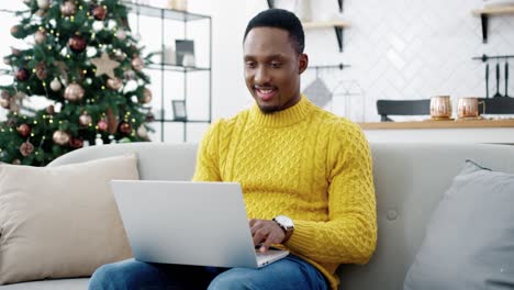 Portrait-Of-Happy-Young-Man-In-Yellow-Sweater-Typing-And-Searching-Internet-On-Laptop-Looking-For-Xmas-Presents-Sales-Online-And-Smiling-In-Cozy-Room-With-Decorated-New-Year-Tree