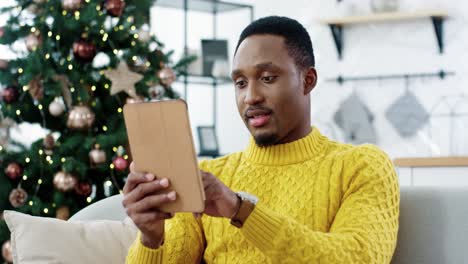 Close-Up-Portrait-Of-Happy-Man-Tapping-On-Tablet-And-Buying-Xmas-Gifts-Online-On-New-Year-Sale-Sitting-Near-Christmas-Tree-In-Room
