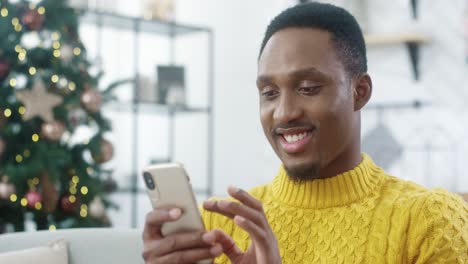 Close-Up-Portrait-Of-Joyful-Man-In-Yellow-Sweater-Sitting-At-Home-With-Christmas-Glowing-Tree-And-Texting-On-Smartphone-Sending-Greetings-And-Wishes-To-Friends-Happy-New-Year-Concept