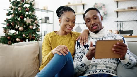 Portrait-Of-Happy-Family-Sitting-On-Sofa-In-Modern-Room-With-Decorated-Glowing-Christmas-Tree-And-Speaking-On-Video-Call-On-Tablet-At-Home-Saying-Holiday-Greetings-To-Friends