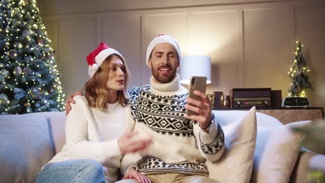 Joyful-Family-Couple-Talking-On-Video-Chat-Online-On-Smartphone-And-Waving-Hands-While-Sitting-At-Home-With-Glowing-Xmas-Tree-Husband-And-Wife-Greeting-Friends-On-Video-Call-New-Year-Concept