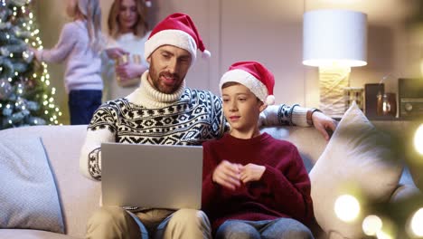 Glücklicher-Vater-Mit-Weihnachtsmütze-Und-Jugendlich-Sohn-Sitzt-In-Der-Nähe-Eines-Leuchtend-Geschmückten-Weihnachtsbaums,-Der-Auf-Einem-Laptop-Tippt-Und-Die-Winterferienzeit-Im-Internet-Verbringt,-Um-Geschenke-Zu-Kaufen