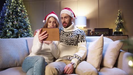 Glückliches-Junges-Ehepaar,-Das-Online-über-Videoanruf-Auf-Dem-Tablet-Spricht,-Während-Es-In-Einem-Gemütlichen-Zimmer-Mit-Weihnachtlich-Geschmücktem,-Leuchtendem-Baum-Sitzt