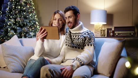 Joyful-Smiling-Family-Couple-Waving-Hands-And-Chatting-On-Video-Call-Online-On-Tablet-With-Friends-While-Sitting-In-Room-With-Christmas-Tree-On-New-Year's-Eve-At-Night-Holiday-Spirit-Concept