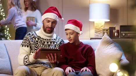 Portrait-Of-Joyful-Dad-With-Teen-Boy-Sitting-In-Cozy-Room-Typing-On-Tablet-Buying-Xmas-Gifts-On-Internet