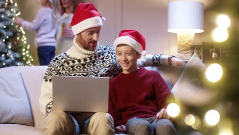 Portrait-Of-Loving-Dad-In-Sana-Hat-With-Teen-Son-Sitting-At-Home-Browsing-On-Laptop-Spending-Holiday-Time-Watching-Xmas-Movie