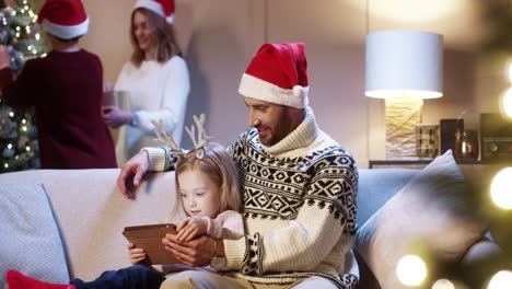 Portrait-Of-Caring-Dad-In-Santa-Hat-With-Cute-Little-Girl-Sitting-In-Room-Tapping-On-Tablet-Buying-Xmas-Gifts-On-Internet-Mom-And-Son-Decorating-Glowing-Christmas-Tree-Family-Holiday-Concept