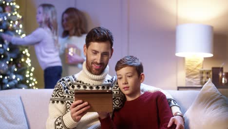 Close-Up-Portrait-Of-Happy-Father-With-Teen-Son-Sitting-In-Room-Tapping-On-Tablet-Choosing-Christmas-Present-On-Internet-While-Mom-And-Cute-Daughter-Decorating-New-Year-Tree-Holidays-Concept