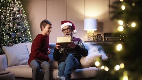 Glücklicher-Süßer-Teenager,-Der-Dem-Glücklichen-Alten-Opa-Weihnachtsgeschenk-Gibt,-Während-Er-In-Einem-Gemütlich-Dekorierten-Zuhause-In-Der-Nähe-Des-Neujahrsbaums-Sitzt