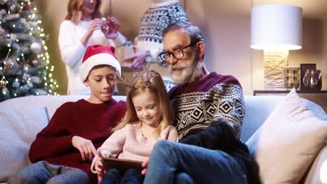 Portrait-Of-Cute-Little-Kids-With-Grandpa-Talking-And-Typing-On-Tablet-Online-Sitting-At-Home-On-Christmas-Eve-While-Parents-Decoration-Xmas-Glowing-Tree-On-Background-Happy-Holidays-Concept