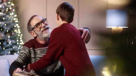 Close-Up-Of-Joyful-Surprised-Old-Grandpa-Receiving-Christmas-Gift-From-Caring-Teen-Boy-While-Sitting-In-Decorated-Room-Near-Glowing-Tree