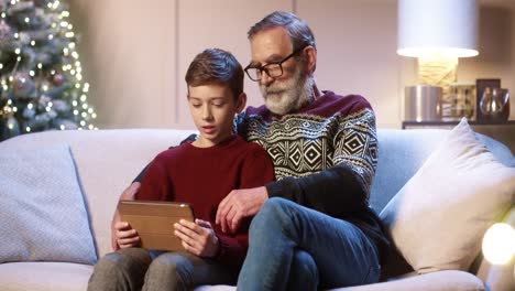 Portrait-Of-Happy-Smiling-Old-Grandpa-With-Teen-Grandchild-Sitting-In-Decorated-Room-Typing-And-Browsing-On-Tablet-While-Chatting-Near-Glowing-New-Year-Tree