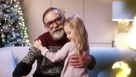 Close-Up-Portrait-Of-Happy-Cute-Little-Girl-Giving-Present-To-Old-Grandpa-While-Sitting-At-Cozy-Decorated-Home-Near-Glowing-New-Year-Tree
