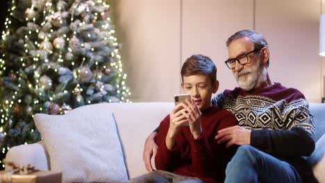 Portrait-Of-Happy-Smiling-Old-Grandpa-With-Teen-Grandchild-Sitting-In-Decorated-Room-And-Taking-Selfie-Photos-On-Smartphone-At-Home-Near-Glowing-New-Year-Tree