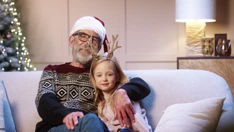 Portrait-Of-Loving-Happy-Senior-Grandpa-In-Glasses-Wearing-Santa-Hat-Spending-Christmas-Together-With-Joyful-Grandchild-Sitting-In-Decorated-Room-Near-Glowing-Xmas-Tree-Embracing-Together