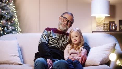 Happy-Caring-Old-Grandfather-In-Glasses-Spending-Time-Together-With-Cute-Little-Granddaughter-Sitting-In-Decorated-House-Near-Glowing-Xmas-Tree-And-Hugging