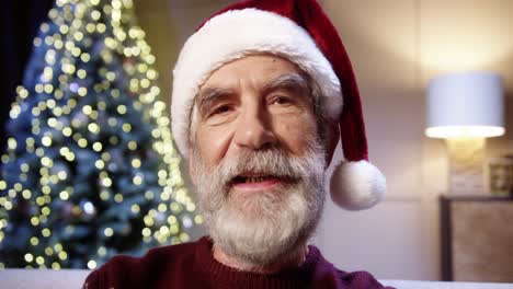 Nahaufnahme-Eines-Glücklichen-Alten-Männlichen-Rentners-Mit-Weihnachtsmütze,-Der-Zu-Hause-Mit-Einem-Glühend-Geschmückten-Weihnachtsbaum-Videochattet