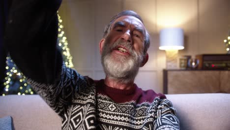Close-Up-Of-Happy-Old-Male-Pensioner-In-Positive-Mood-Video-Chatting-Near-Glowing-Decorated-Xmas-Tree