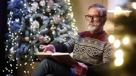 Portrait-Of-Old-Nice-Male-Pensioner-Grandpa-In-Glasses-And-Christmas-Sweater-Reading-Book-While-Sitting-In-Decorated-Room-Near-Glowing-Christmas-Tree