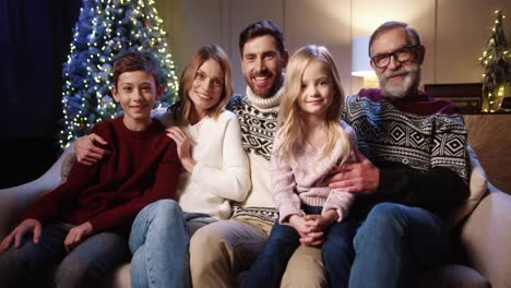 Close-Up-Of-Cheerful-Family-Sitting-Together-At-Cozy-Home-Near-Decorated-Glowing-Xmas-Tree-Smiling-And-Looking-At-Camera-On-Christmas-Eve