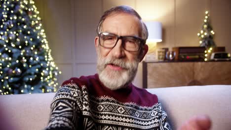 Close-Up-Of-Cheerful-Smiling-Old-Male-Pensioner-In-Glasses-Chatting-On-Video-Call-In-Room-With-Glowing-Decorated-Xmas-Tree