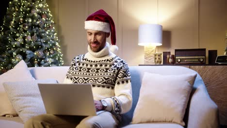 Portrait-Of-Happy-Handsome-Male-In-Santa-Hat-Sitting-In-Cozy-Room-With-Xmas-Tree-And-Typing-On-Laptop-Looking-Away