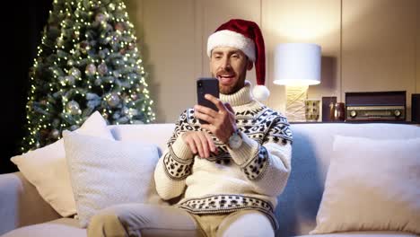 Fröhlicher-Gutaussehender-Mann-Mit-Weihnachtsmütze-Sitzt-In-Einem-Gemütlichen-Zimmer-Mit-Weihnachtsbaum-Und-Video-Chat-Auf-Dem-Smartphone-Und-Gratuliert-Mit-Feiertagen