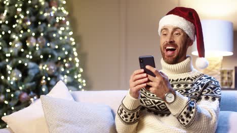 Nahaufnahme-Porträt-Eines-Fröhlichen,-Gutaussehenden-Mannes-Mit-Weihnachtsmütze,-Der-In-Einem-Gemütlichen-Zimmer-In-Der-Nähe-Des-Weihnachtsbaums-Sitzt-Und-Mit-überraschtem-Gesicht-Auf-Dem-Smartphone-Tippt,-Um-Die-Weihnachtslotterie-Zu-Gewinnen