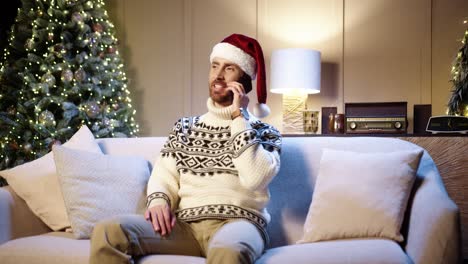 Fröhlicher-Gutaussehender-Mann-Mit-Weihnachtsmütze,-Der-Zu-Hause-Mit-Weihnachtsbaum-Sitzt-Und-Auf-Dem-Smartphone-Chattet-Und-Freunden-Mit-Feiertagen-Gratuliert