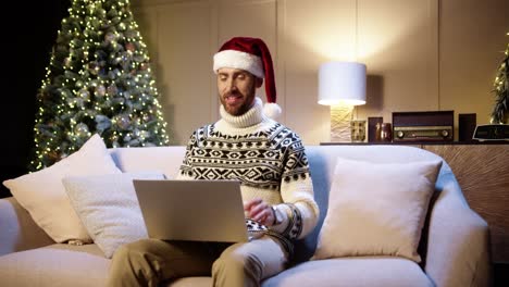 Portrait-Of-Happy-Young-Man-In-Santa-Hat-Sitting-In-Decorated-Room-Near-Glowing-Xmas-Tree-Speaking-On-Video-Chat-On-Laptop